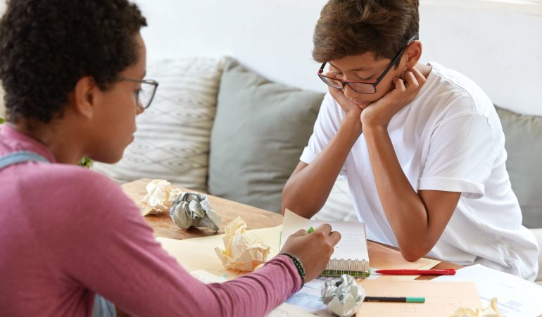 Back view of experienced Afro woman explains school education content to male hipster, indicates at information written in notepad, pose in cozy place. Assistance, cooperation and tutoring concept