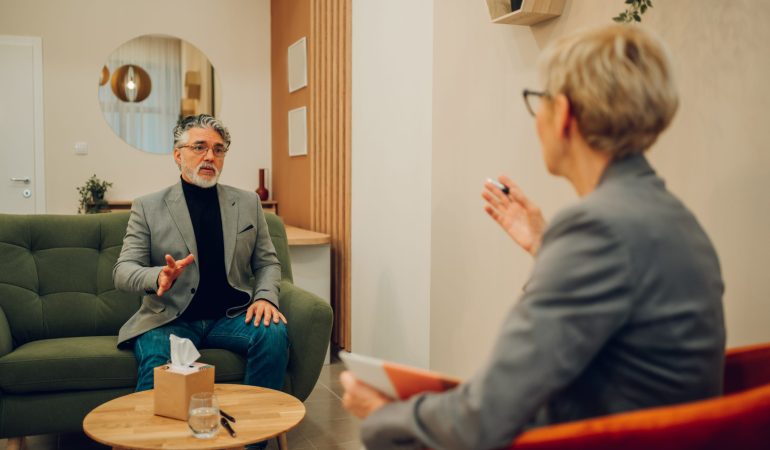 A senior man is sitting in a doctor's office with a shrink and discussing problems during the session. A psychotherapist is giving a man a supportive talk while holding a textbook.
