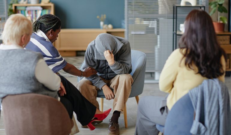 Group of people supporting distressed teenager sitting with hood over head in therapy session, showing empathy and comfort in mental health support setting